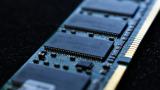 Macro close-up of a computer RAM module, showing the blue circuit board and metallic connectors in sharp detail with a shallow depth-of-field blur.

